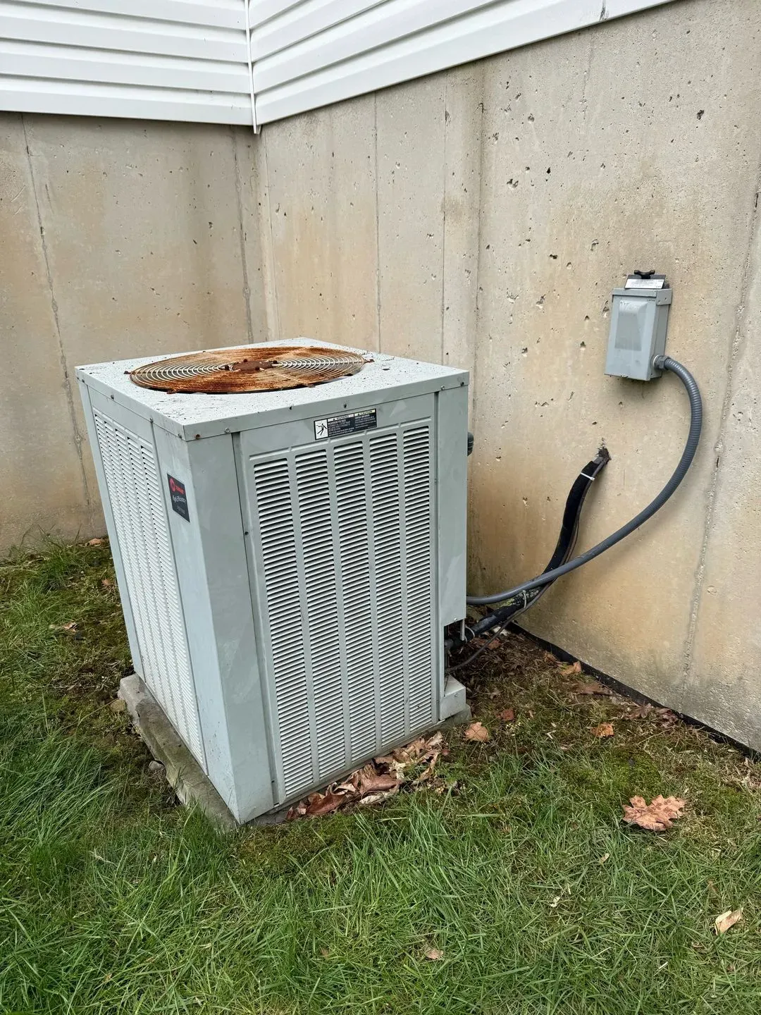 An outdoor air conditioning unit with a rusty top, mounted near a concrete wall.