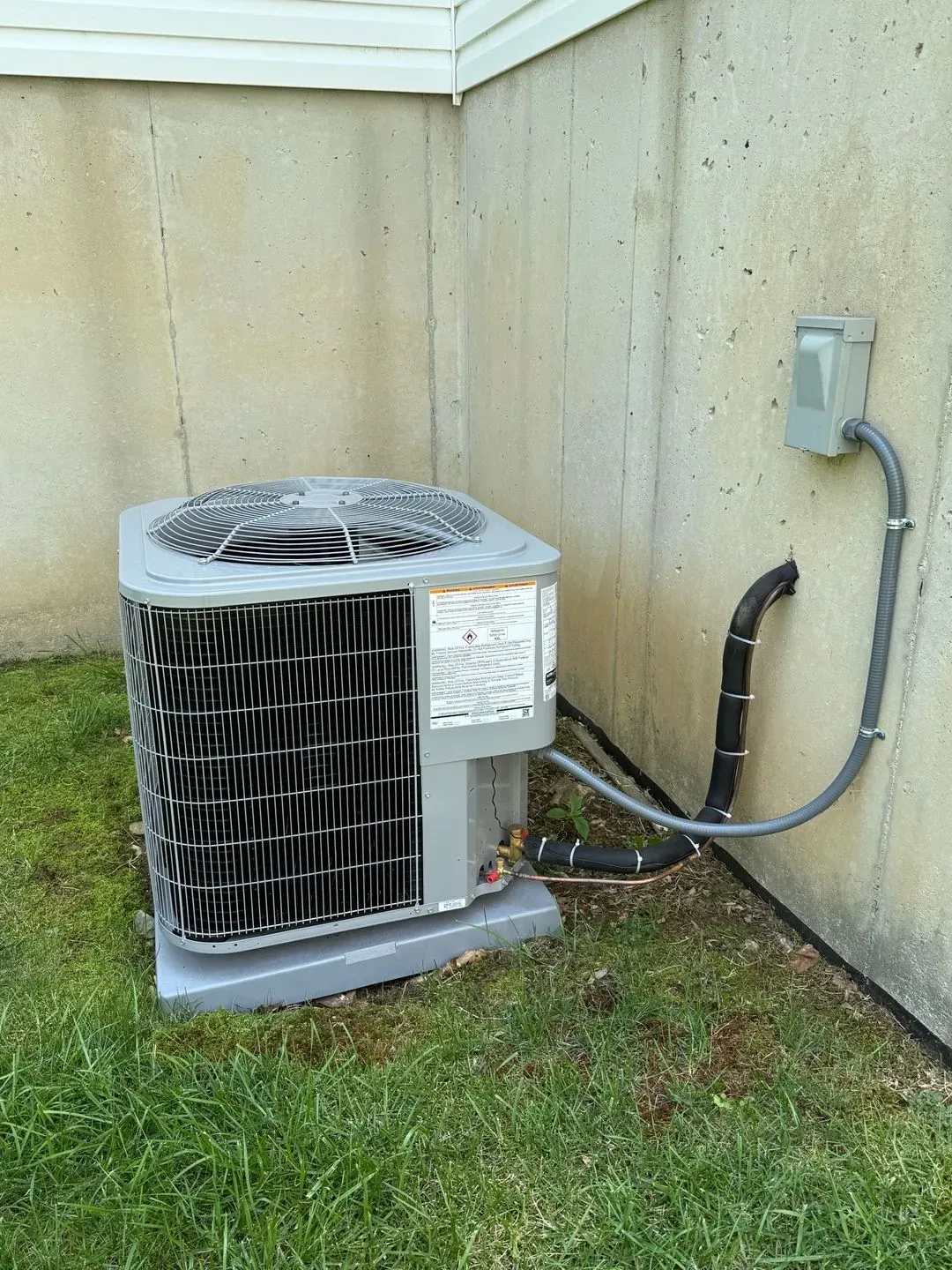 Air conditioning unit next to concrete wall with electrical conduit and tubing on green grass.
