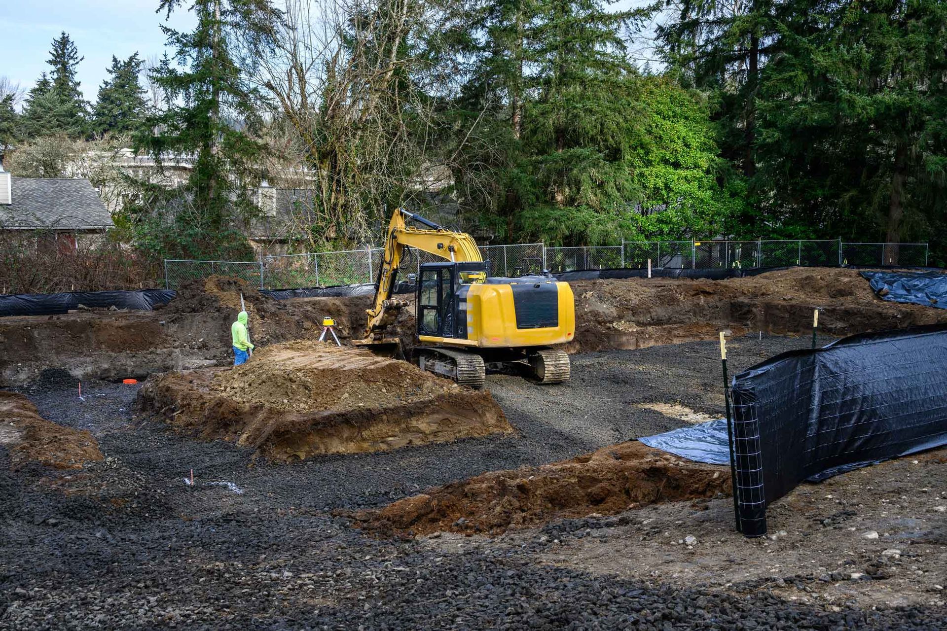 Construction site with yellow excavator and worker. Dirt mounds, black fabric, and trees in background.