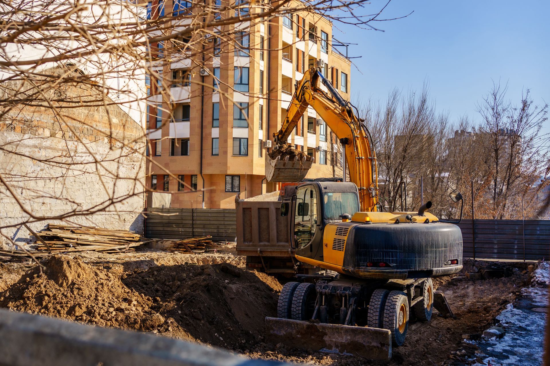 Excavator loads dirt into a dump truck at a construction site near a tall building.