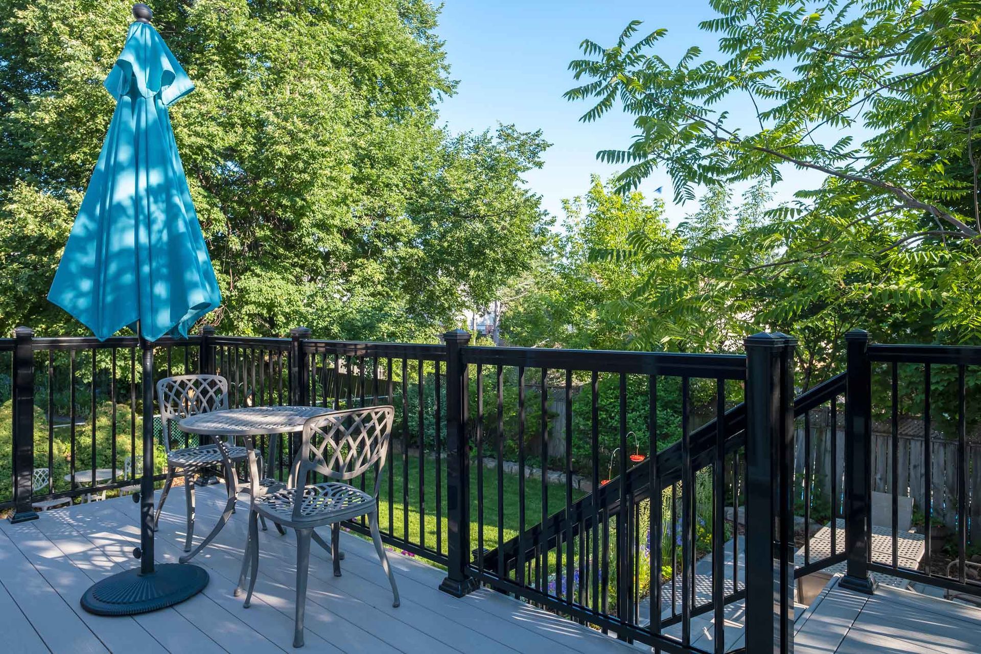 Outdoor deck with table, chairs, and blue umbrella, overlooking a green yard and trees.