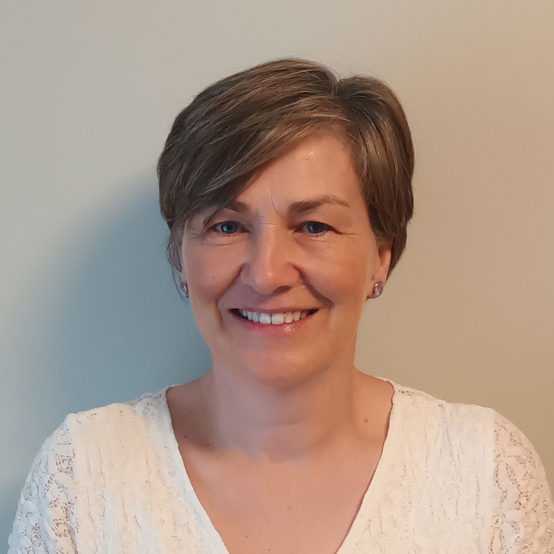 Woman with short, light brown hair smiles, wearing a white lace top, against a plain wall.