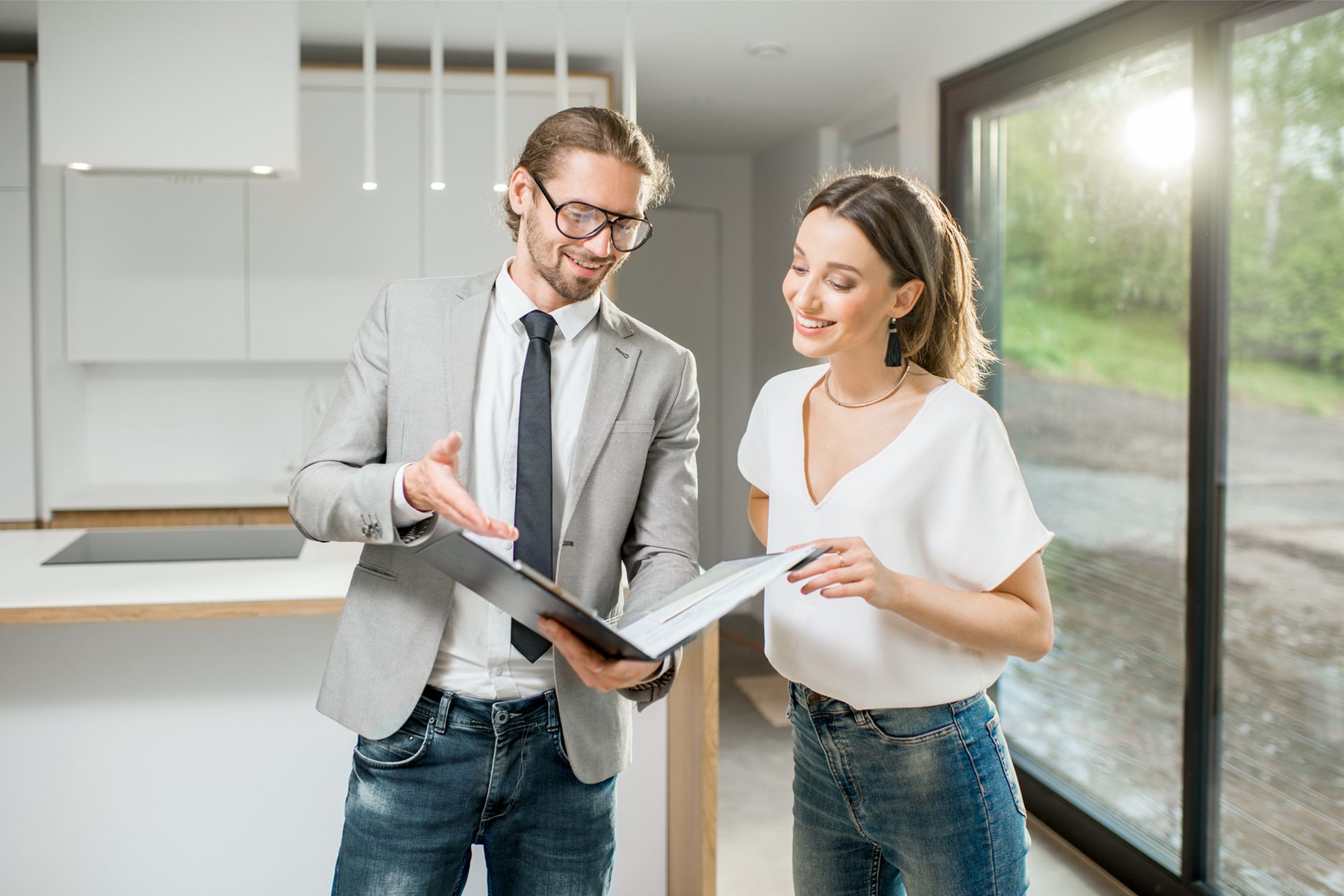 Real estate agent showing a document to a woman in a modern kitchen with large windows.