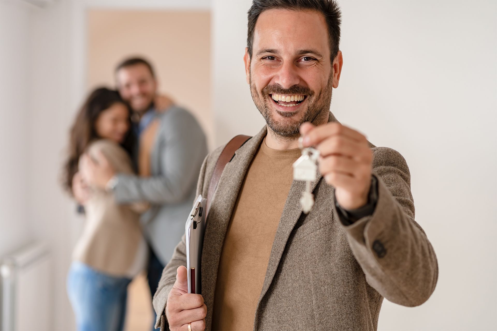 Real estate agent smiling, holding keys; happy couple embraces in background.