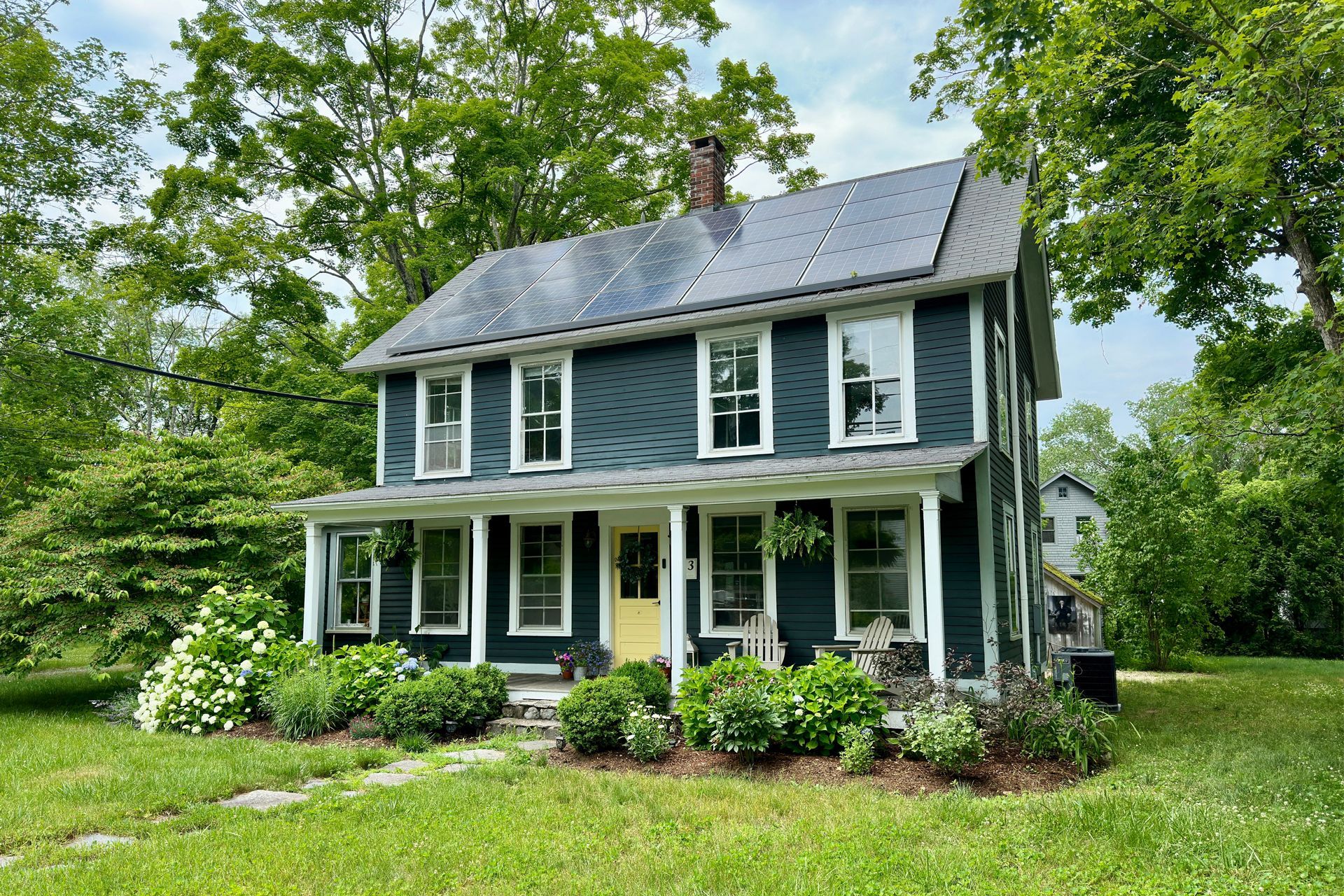 Two-story teal house with white trim, solar panels, and yellow door surrounded by greenery.