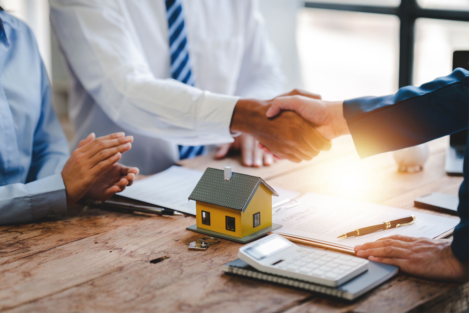 People shaking hands over a desk, miniature house, calculator, and paperwork; real estate deal.