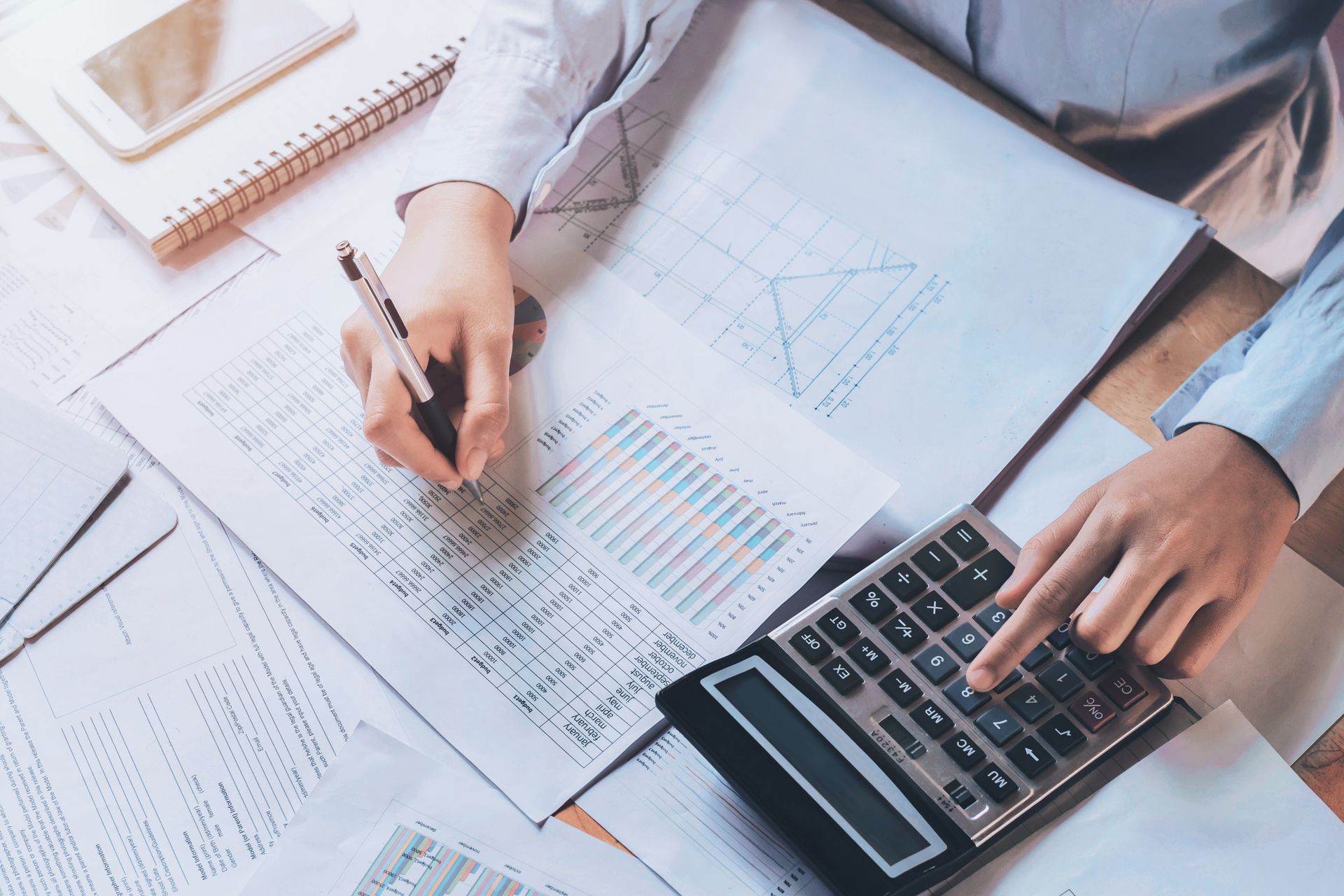 Hands calculating on a calculator beside financial charts and a pen on a desk.