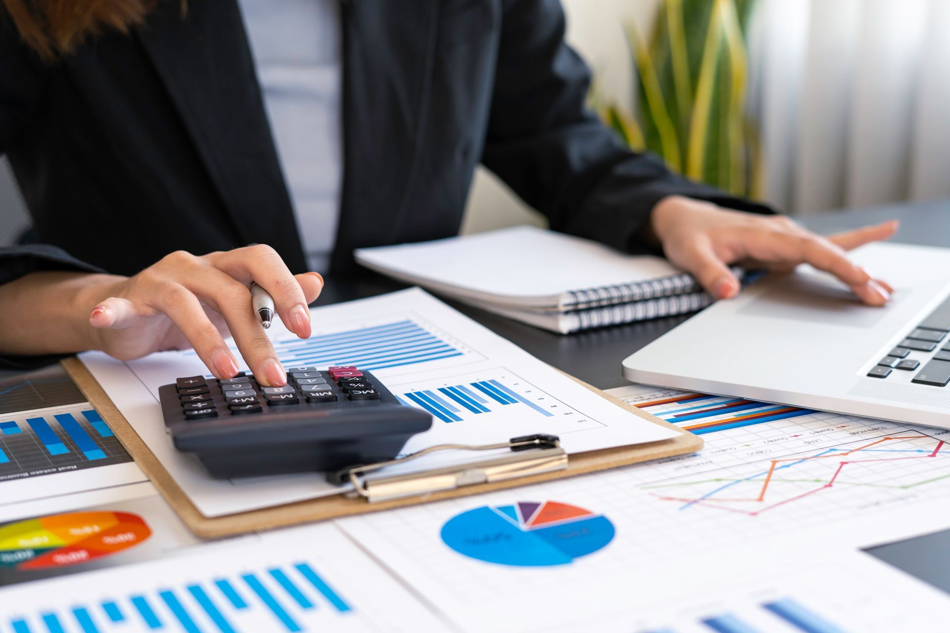 Person using a calculator over charts and graphs beside a laptop on a desk