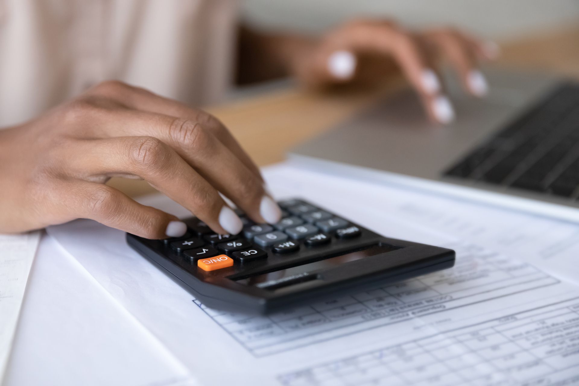 Hands using a calculator on paperwork beside a laptop