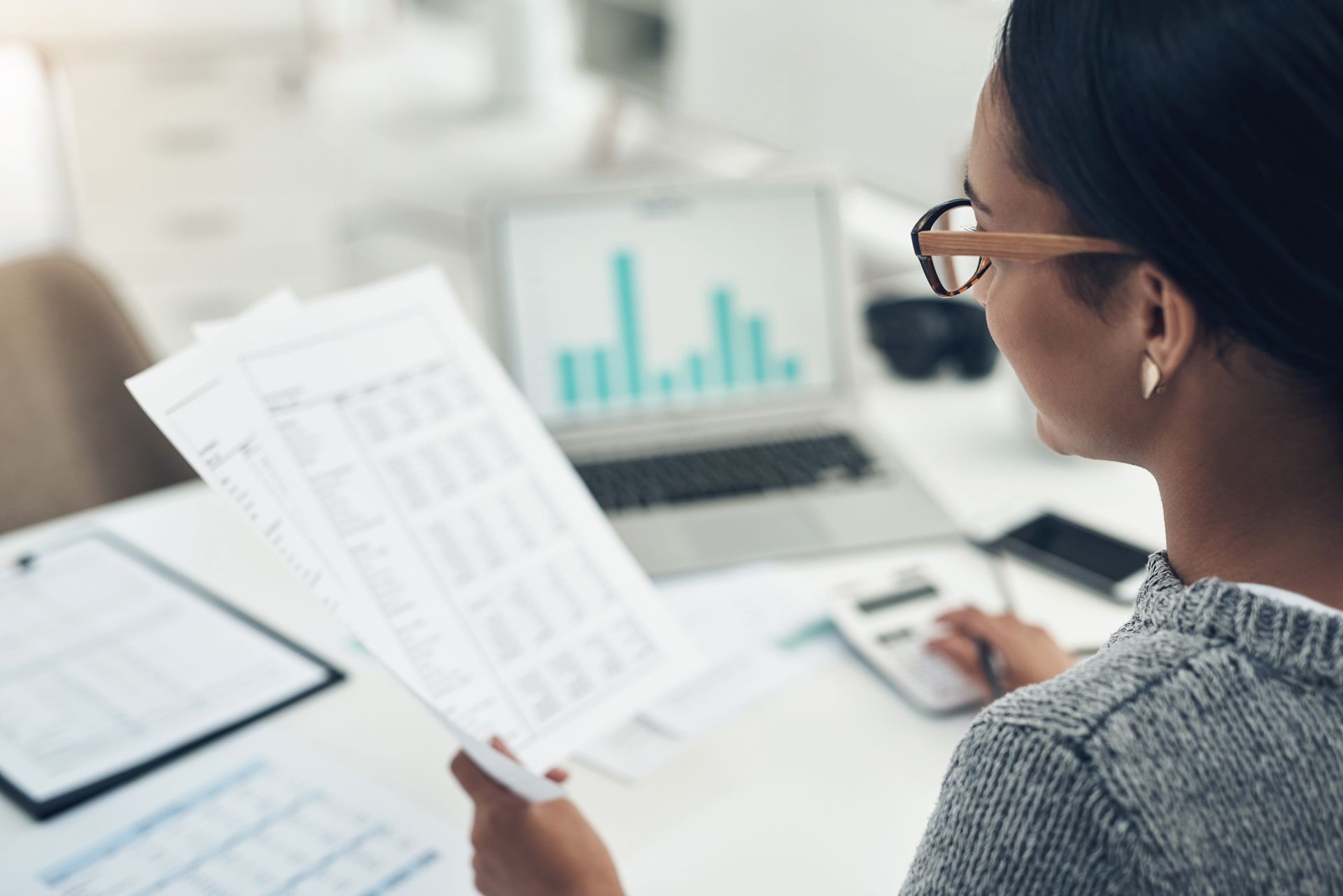 Person reviewing printed charts at a desk with a monitor showing analytics graphs