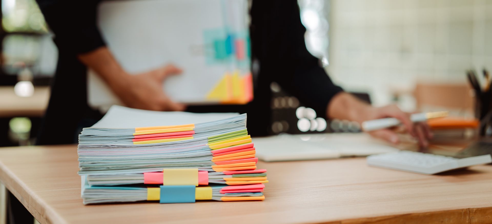 Stack of colorful sticky notes and papers on a desk with people working in the background