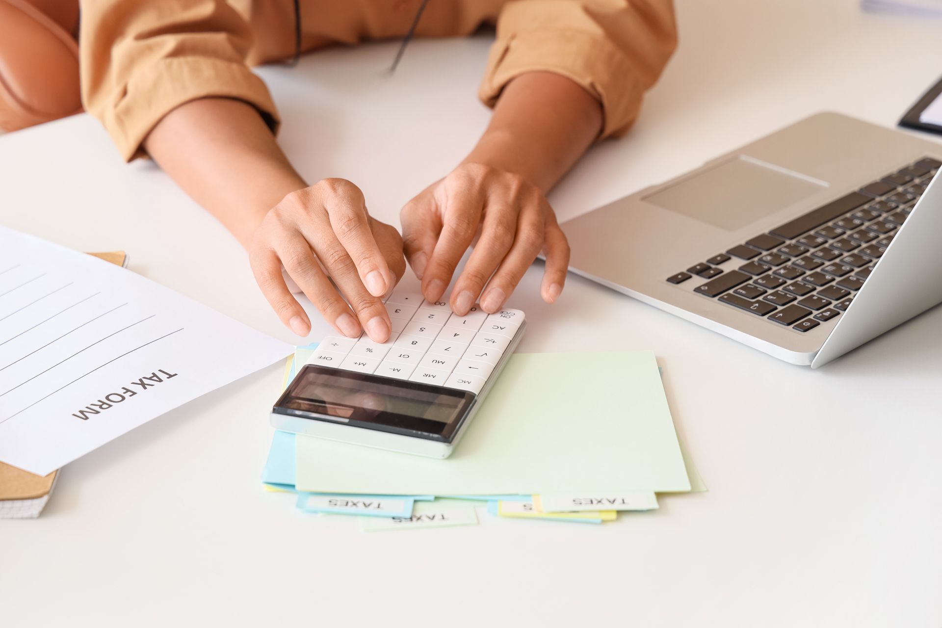 Hands typing on a calculator beside a laptop and paperwork on a white desk