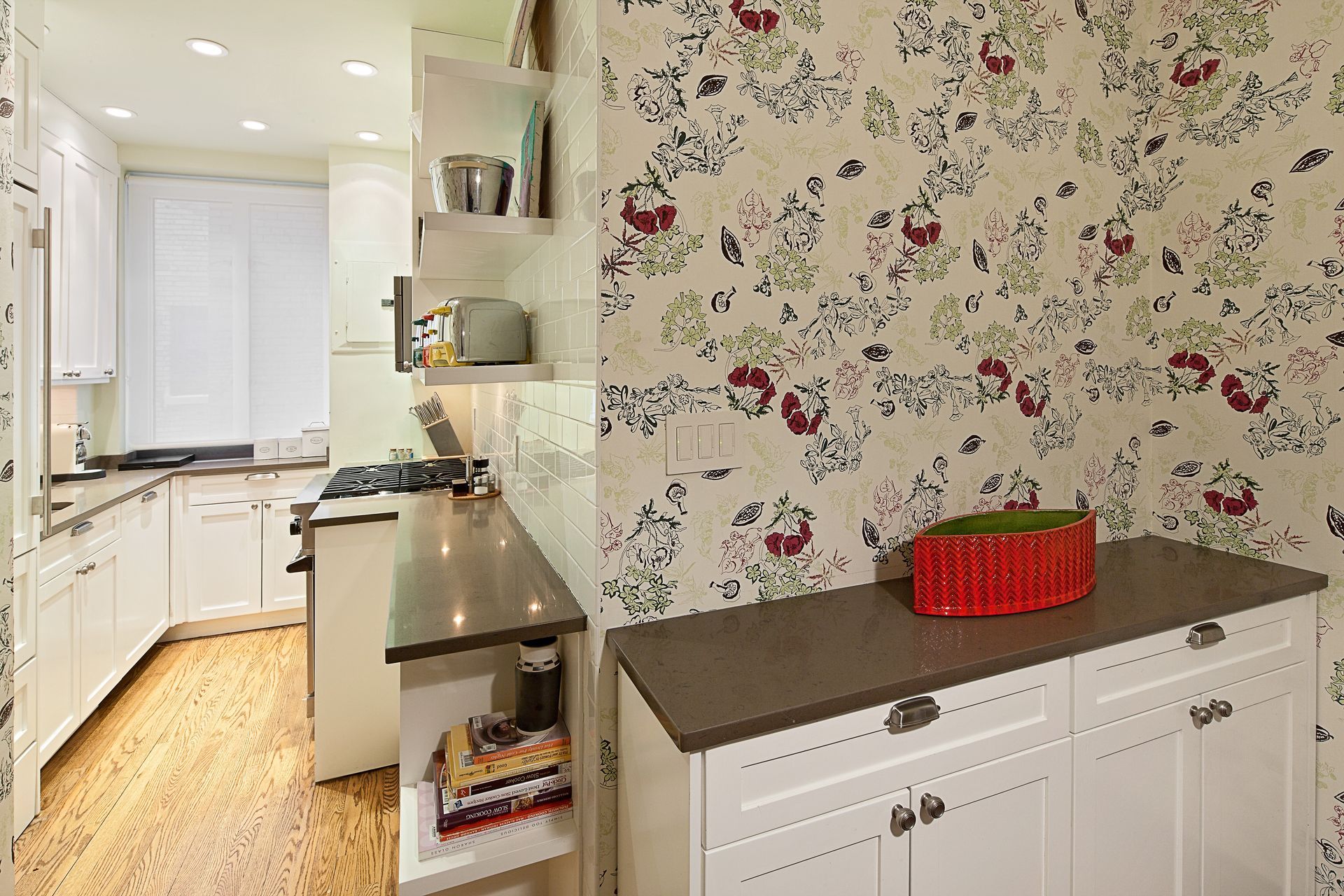 A kitchen with white cabinets, wood floors, a floral wallpaper accent wall, and a low white cabinet with a red bowl.