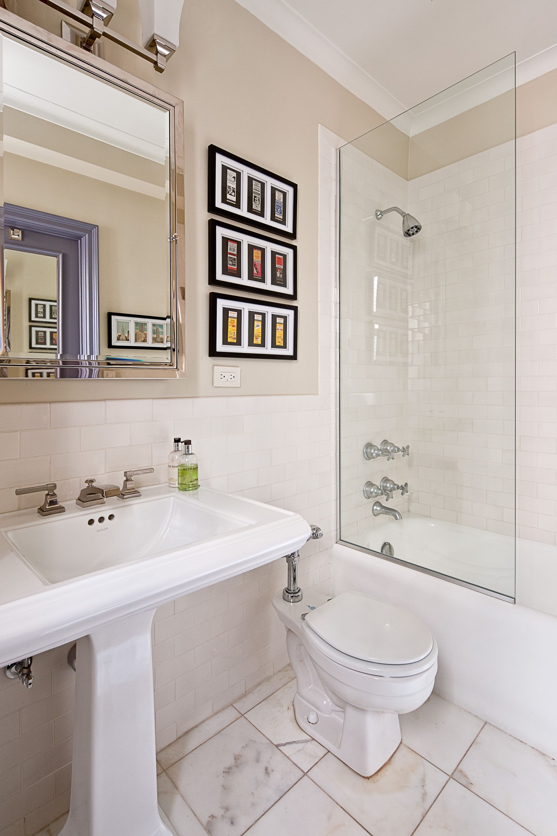 A clean, neutral-toned bathroom with a pedestal sink, framed wall art, and a glass-enclosed shower over a bathtub.