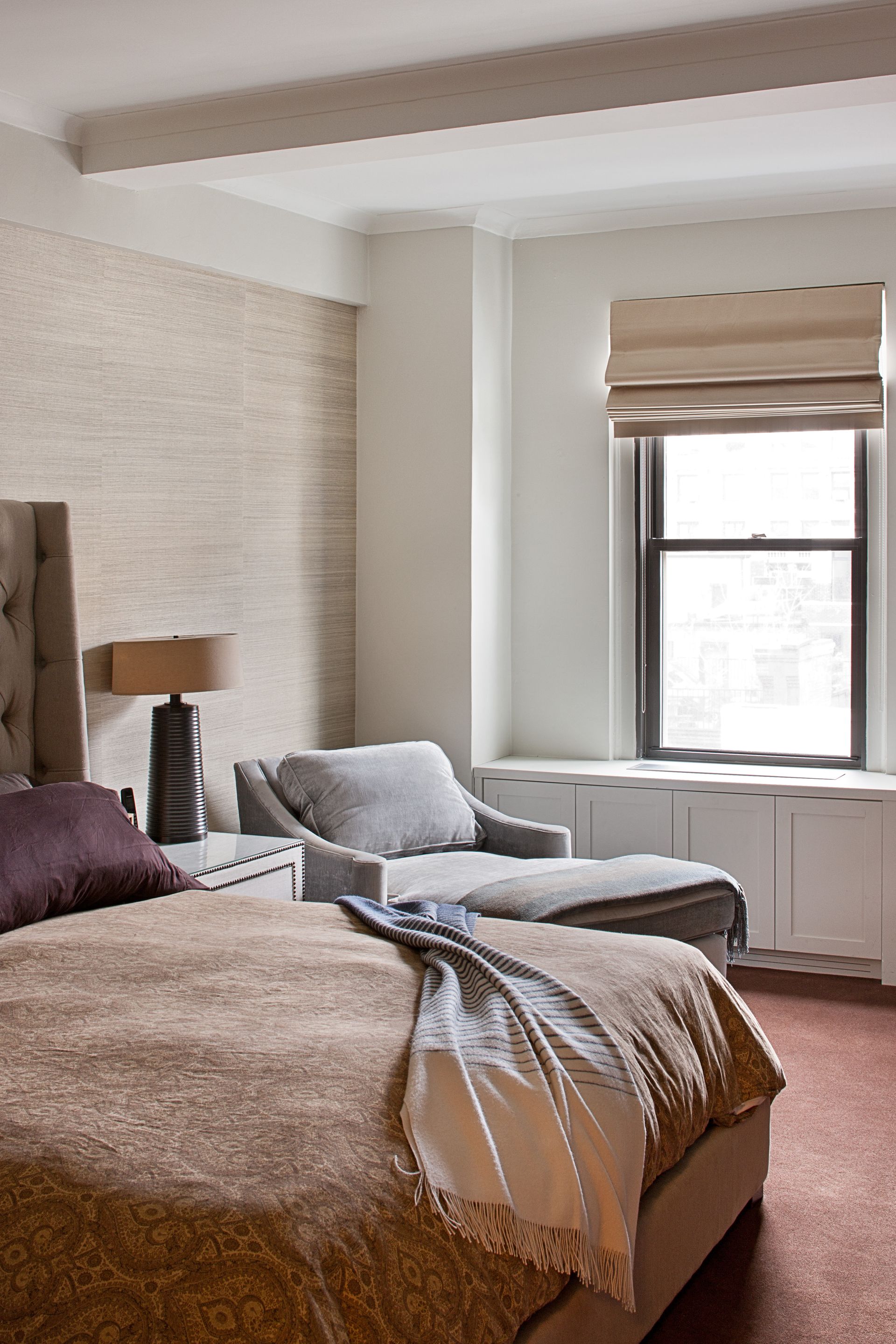 A neutral-toned bedroom with a bed featuring a textured throw, a side lamp, and a chaise longue near a window.
