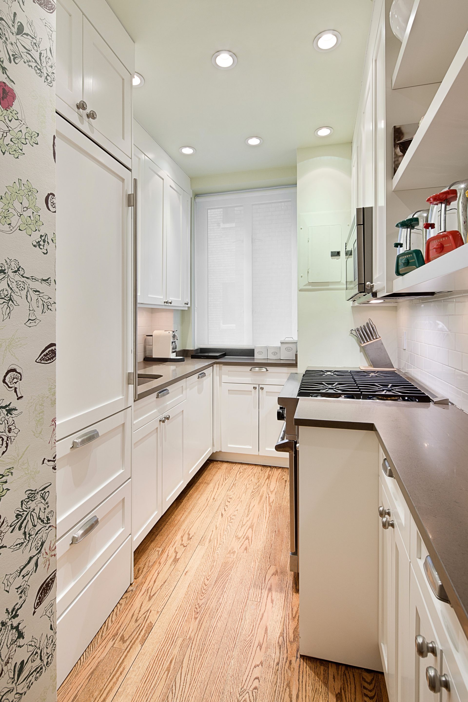 Narrow galley kitchen with white cabinets, dark countertops, light wood flooring, and patterned wallpaper.