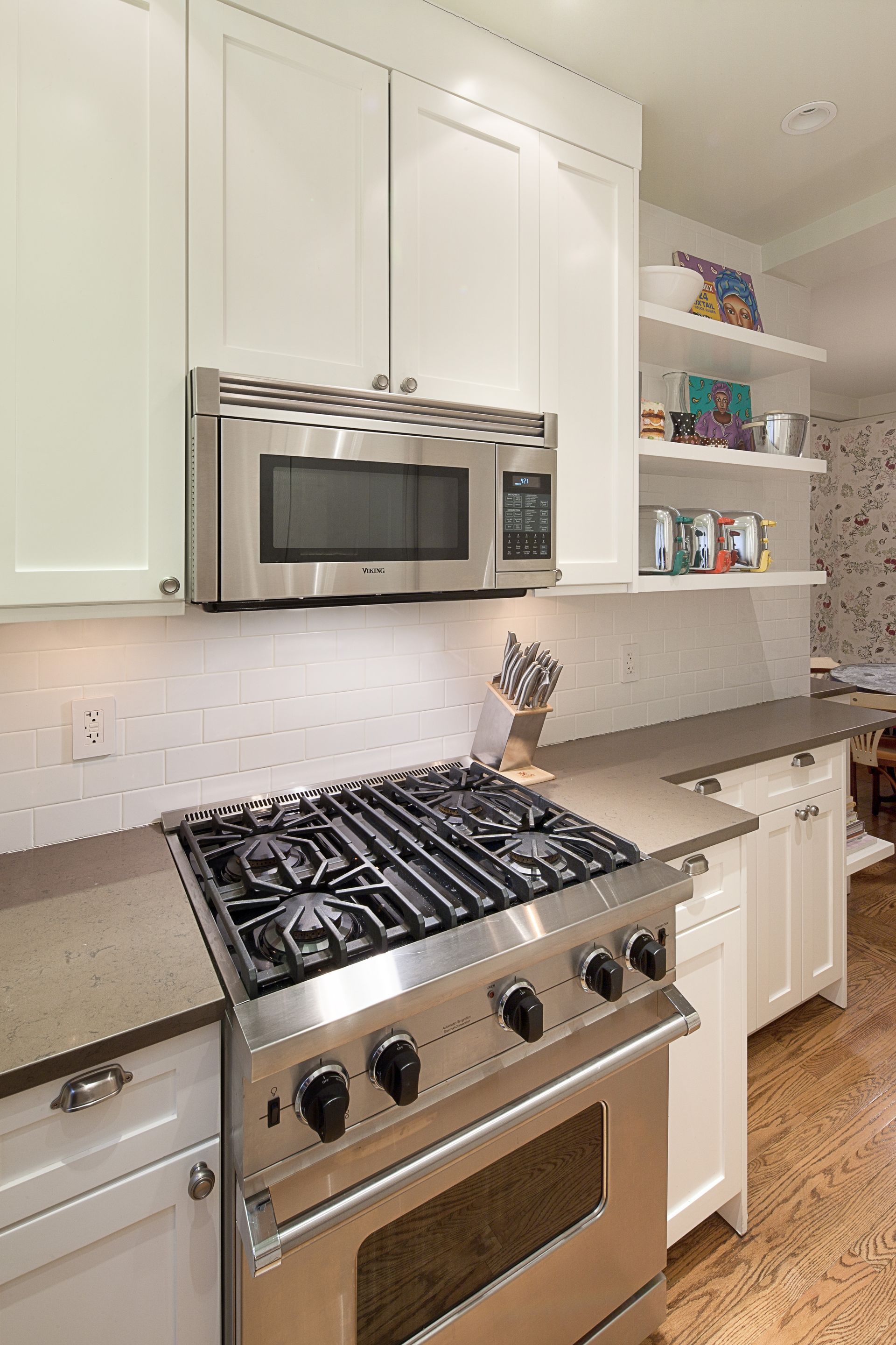 Stainless steel gas range and microwave set into white cabinets with grey countertops and open shelving in a kitchen.