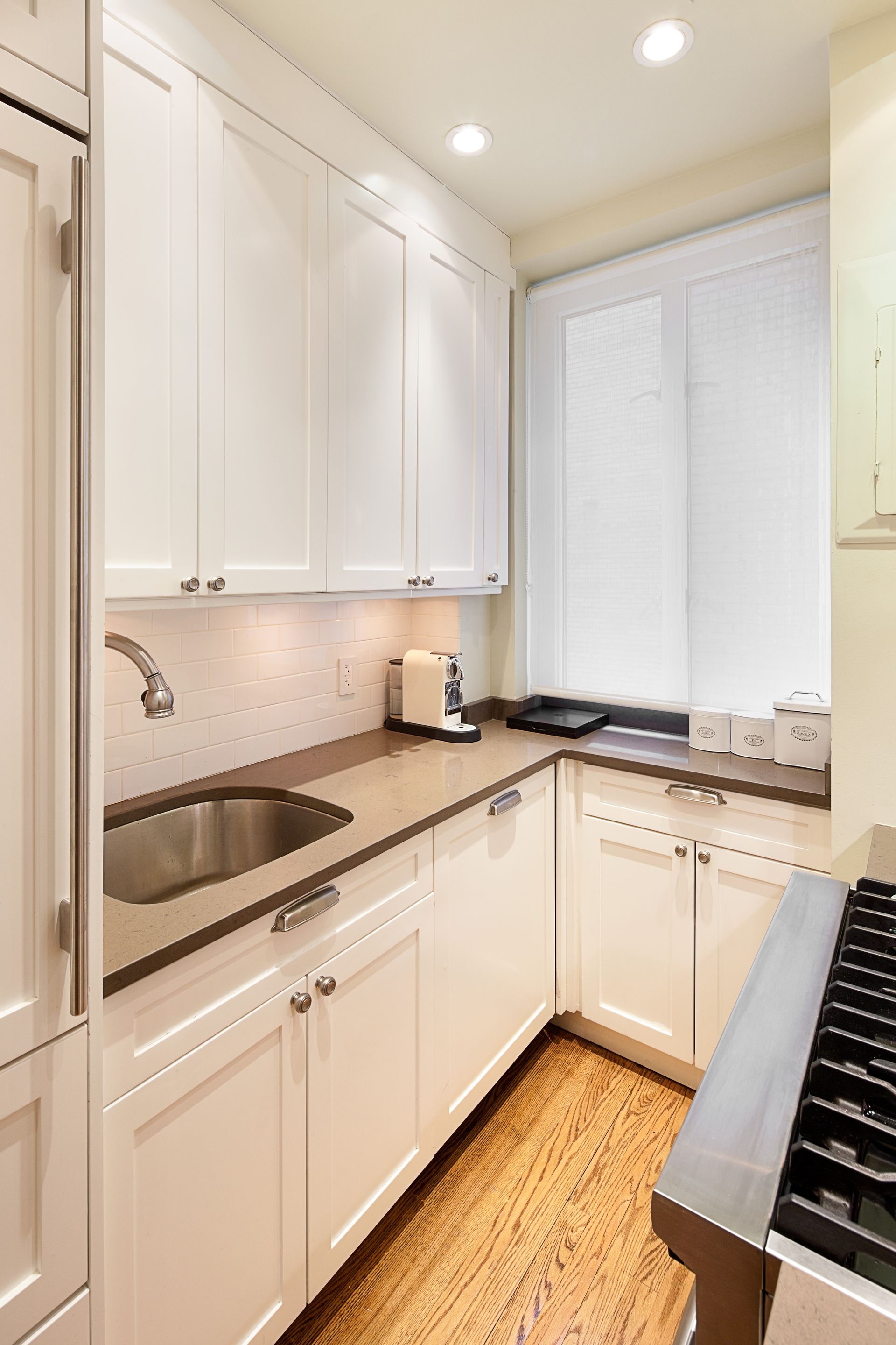 A small, narrow kitchen with white cabinets, a stainless steel sink, beige countertops, and wood-patterned flooring.