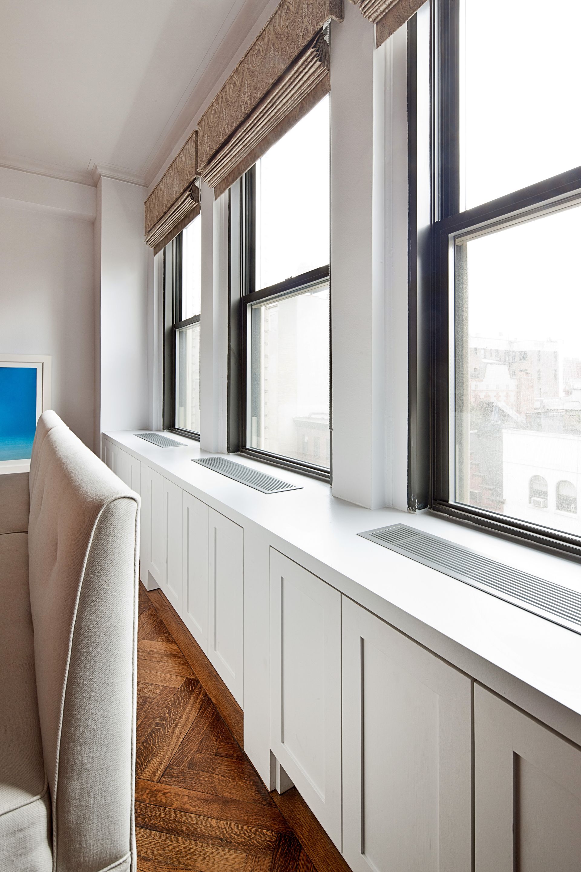 Modern windows with white built-in radiator covers below, set in a room with hardwood floors and light-colored furniture.