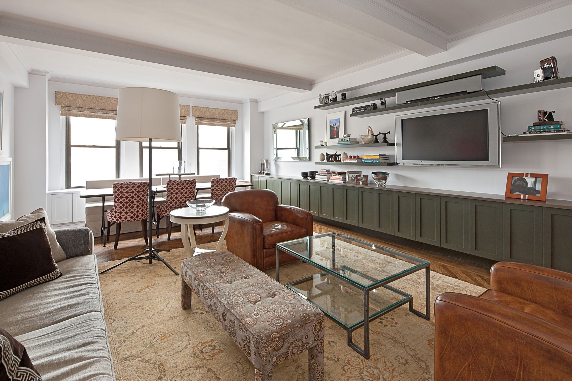 A living room with a brown leather armchair, glass coffee table, sofa, and a large wall-mounted TV over green cabinets.