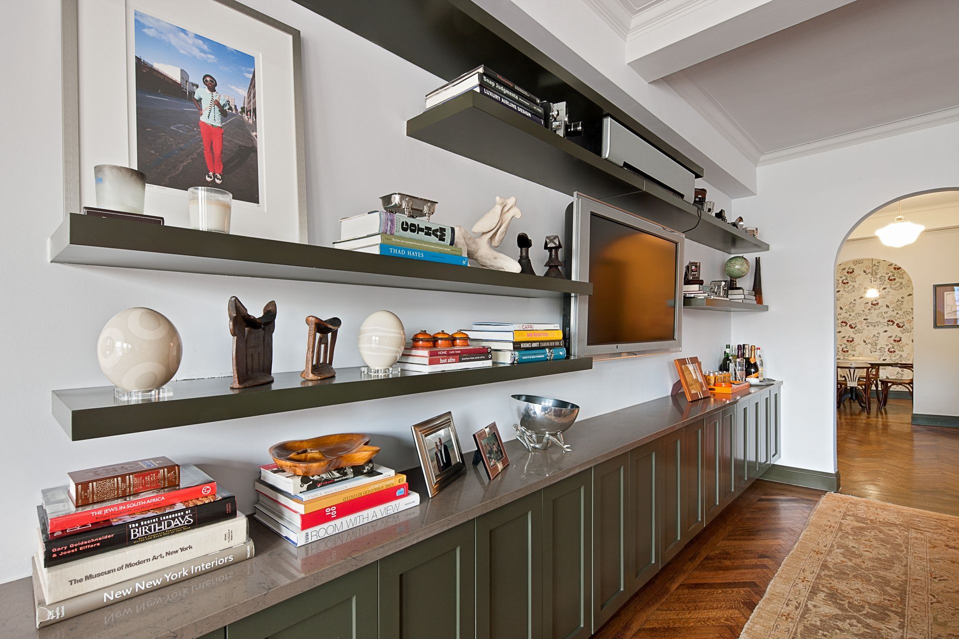 A living room wall with dark green built-in cabinetry, floating shelves filled with books and decor, and a wall-mounted TV.