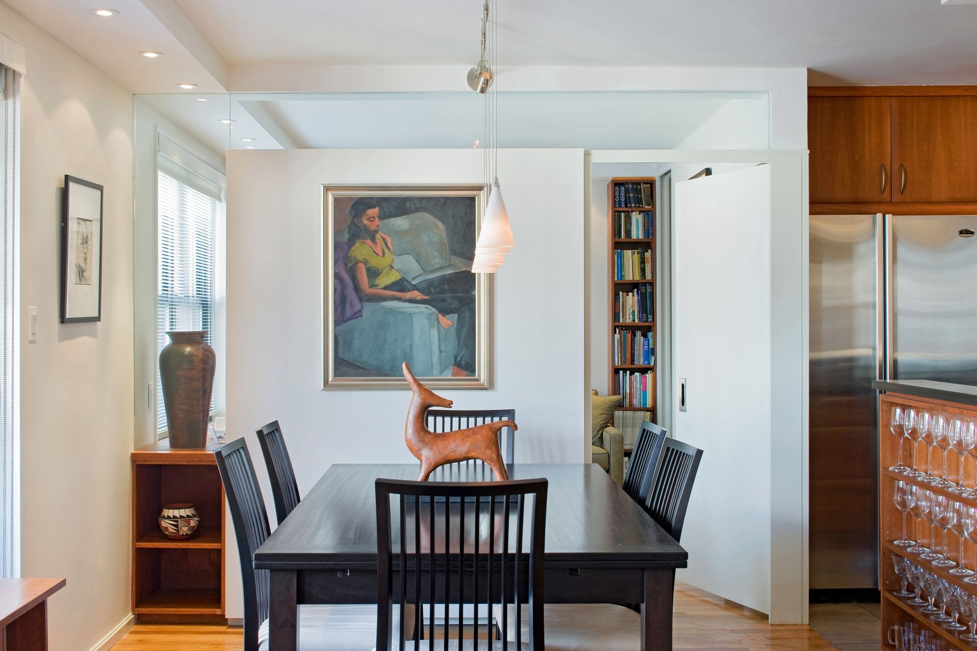 A dining room with a dark wooden table, black chairs, a wall painting, and an adjacent kitchen with wooden cabinets.