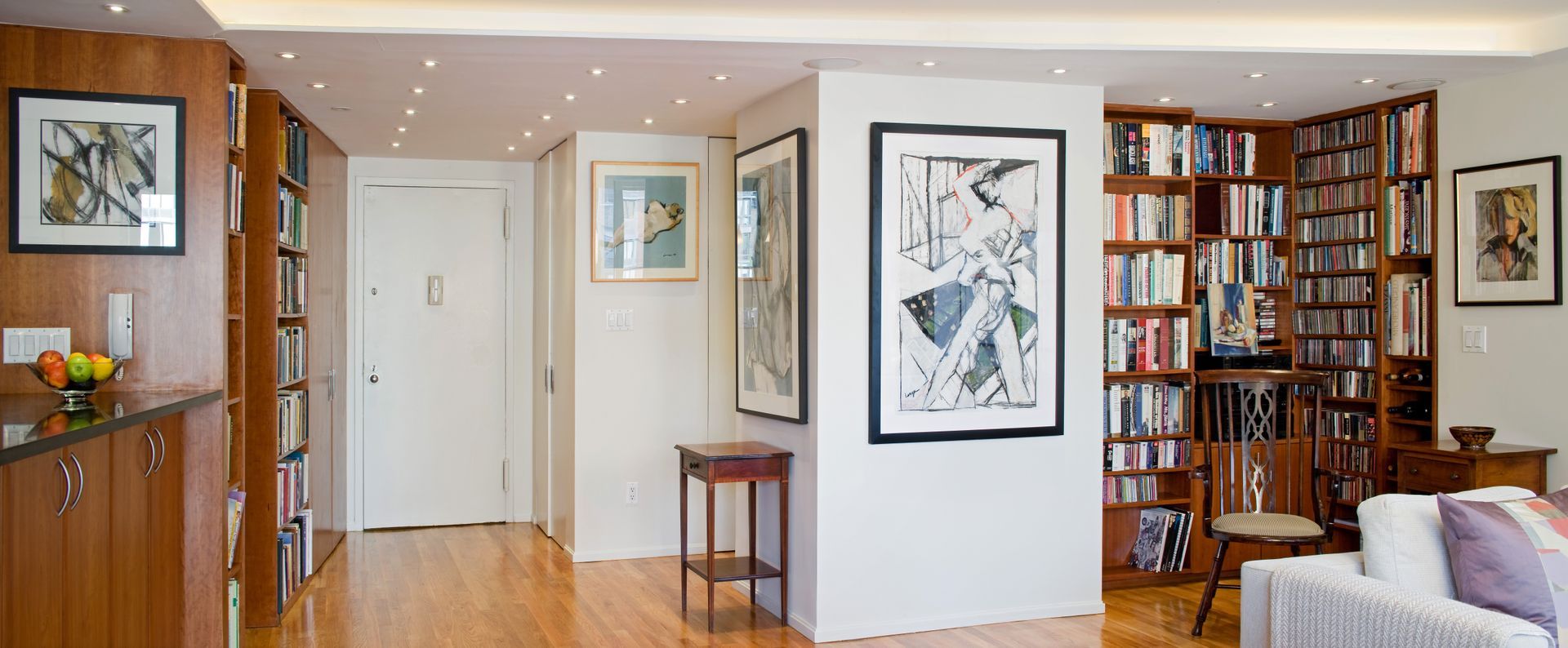 A bright interior living space featuring wooden bookshelves, white walls, framed artwork, and a white doorway.