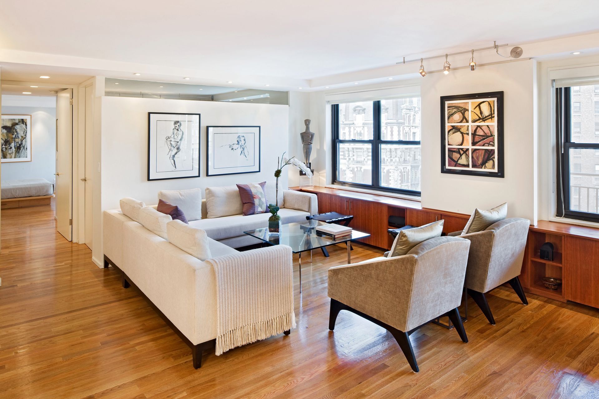 A modern living room with a white sofa, two chairs, a glass coffee table, and framed art on white walls over hardwood.