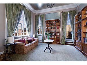 A bright, elegant room with high ceilings, tall windows with green curtains, a patterned sofa, a small table, and shelves.