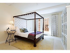 A bedroom with a dark wood canopy bed, light-patterned drapes, a side table with a lamp, and a tall cabinet on a rug.
