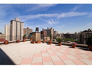 A sunny rooftop terrace with potted plants overlooking a city skyline of tall apartment buildings under a blue sky.