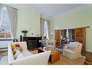 A living room with pale green walls, a white sofa, two armchairs, a wooden coffee table, and large windows with curtains.