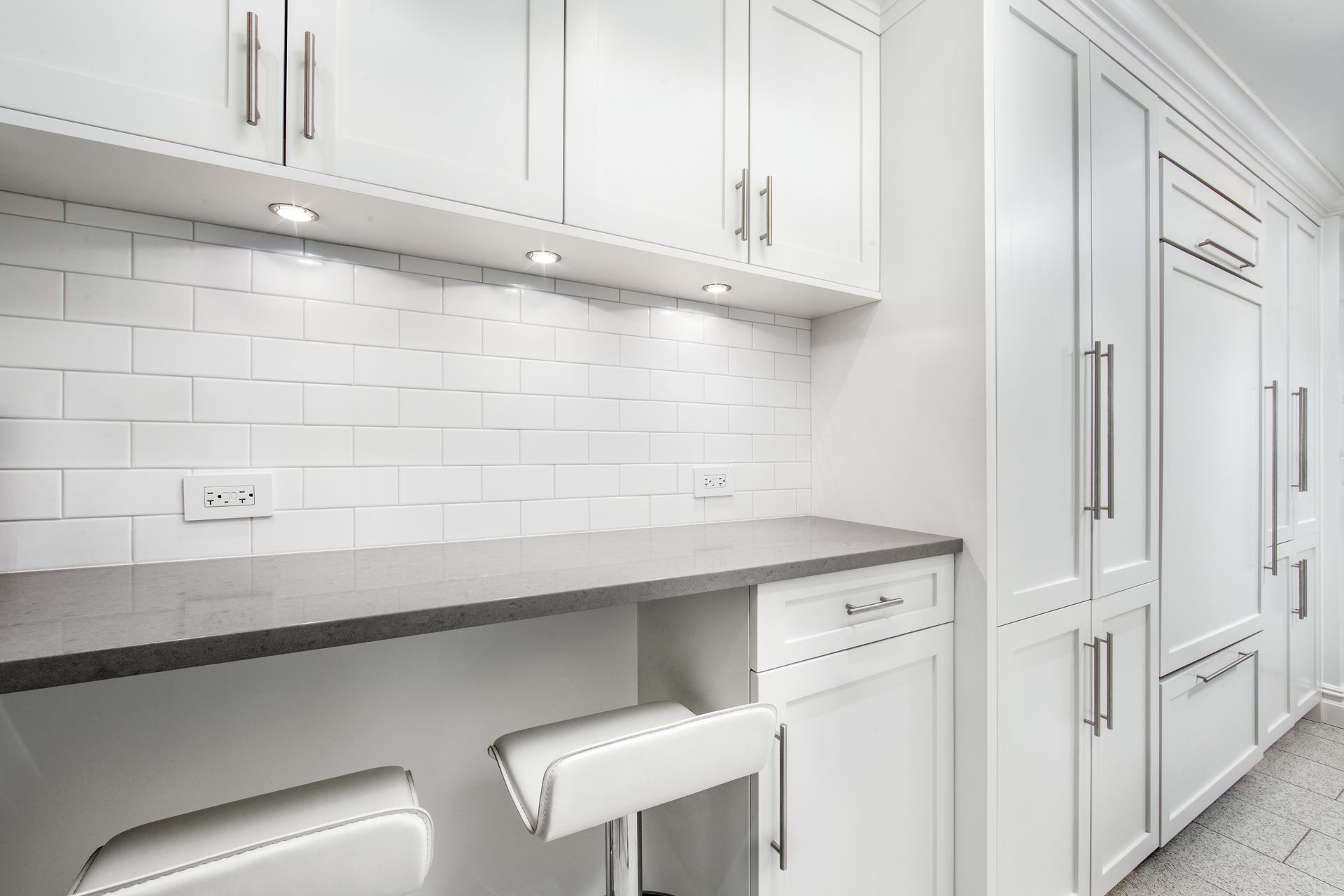 A modern kitchen featuring white cabinets, a dark gray countertop, white subway tile backsplash, and two white stools.