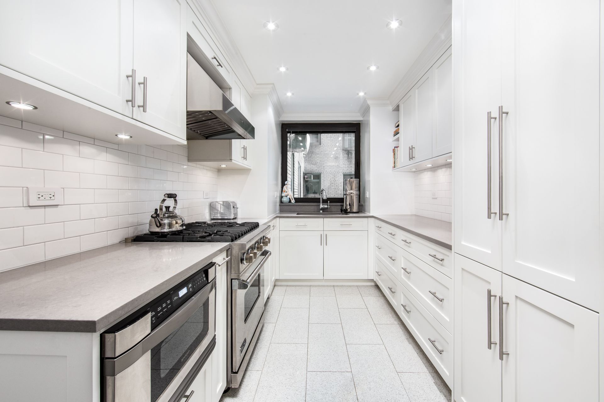 A bright, narrow galley kitchen with white cabinets, stainless steel appliances, grey countertops, and light tiled floors.