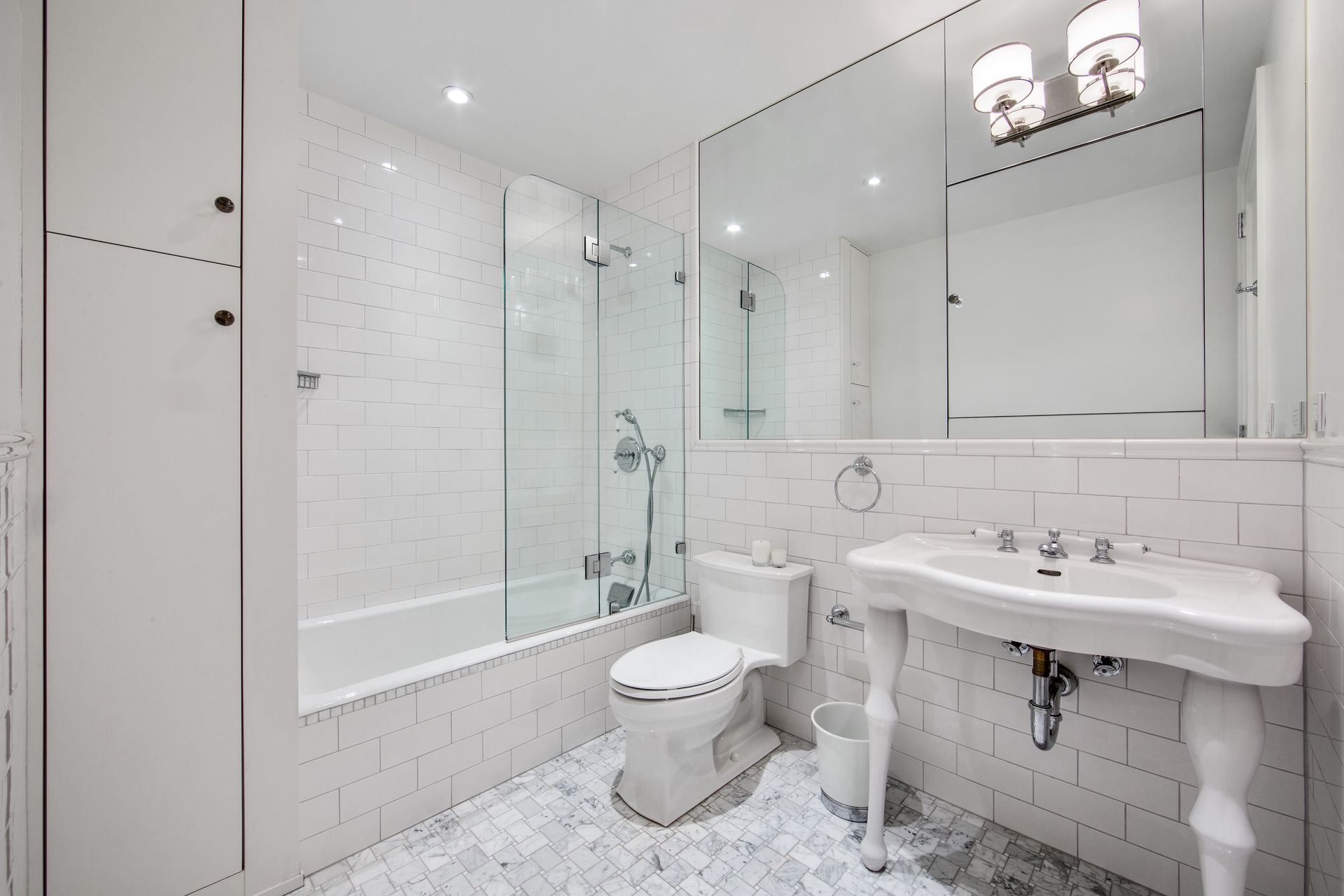 A bright, modern white bathroom featuring a bathtub with a glass screen, a pedestal sink, a toilet, and tiled surfaces.