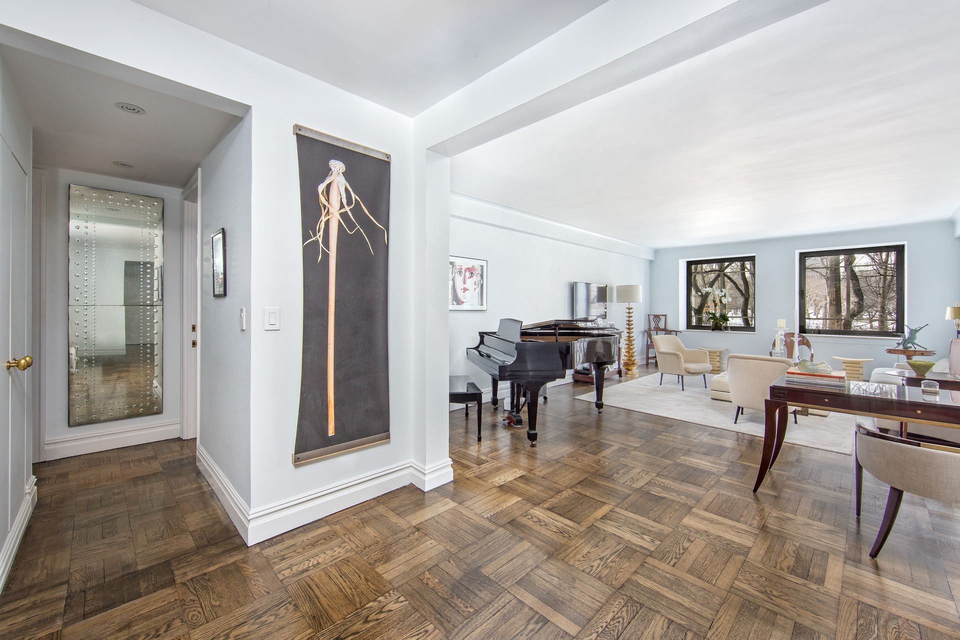 A brightly lit living room with parquet floors, a grand piano, and two windows overlooking trees.