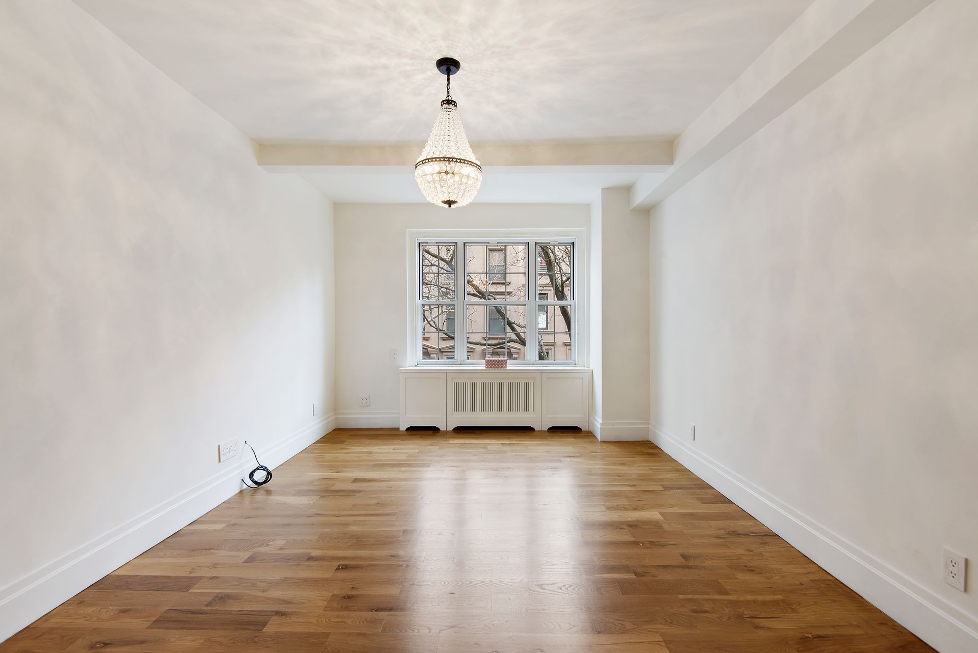 A bright, empty living room with white walls, wooden floors, a crystal chandelier, and a window with a radiator cover.
