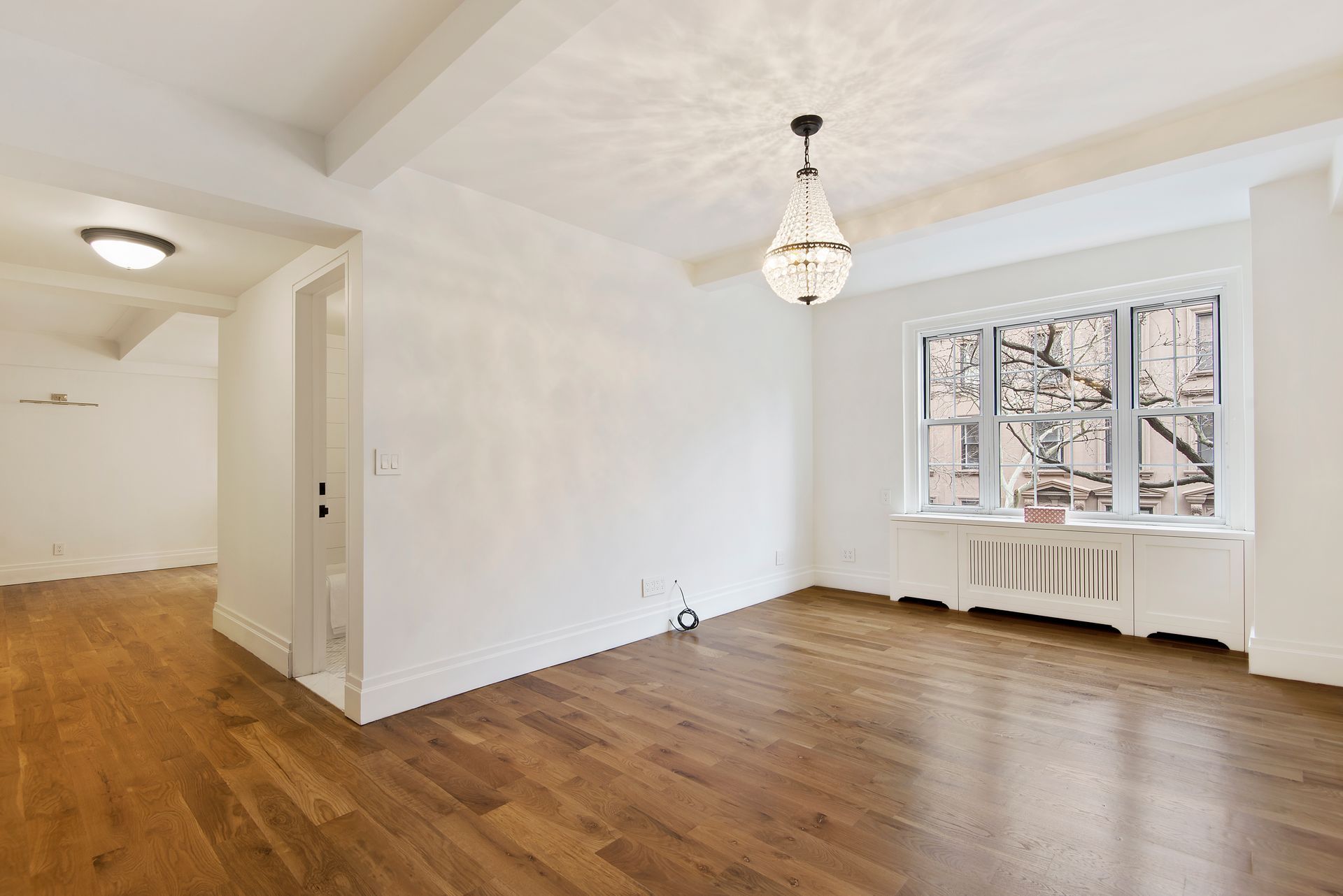 A bright, empty living room with light-colored walls, hardwood floors, a crystal chandelier, and a large window.
