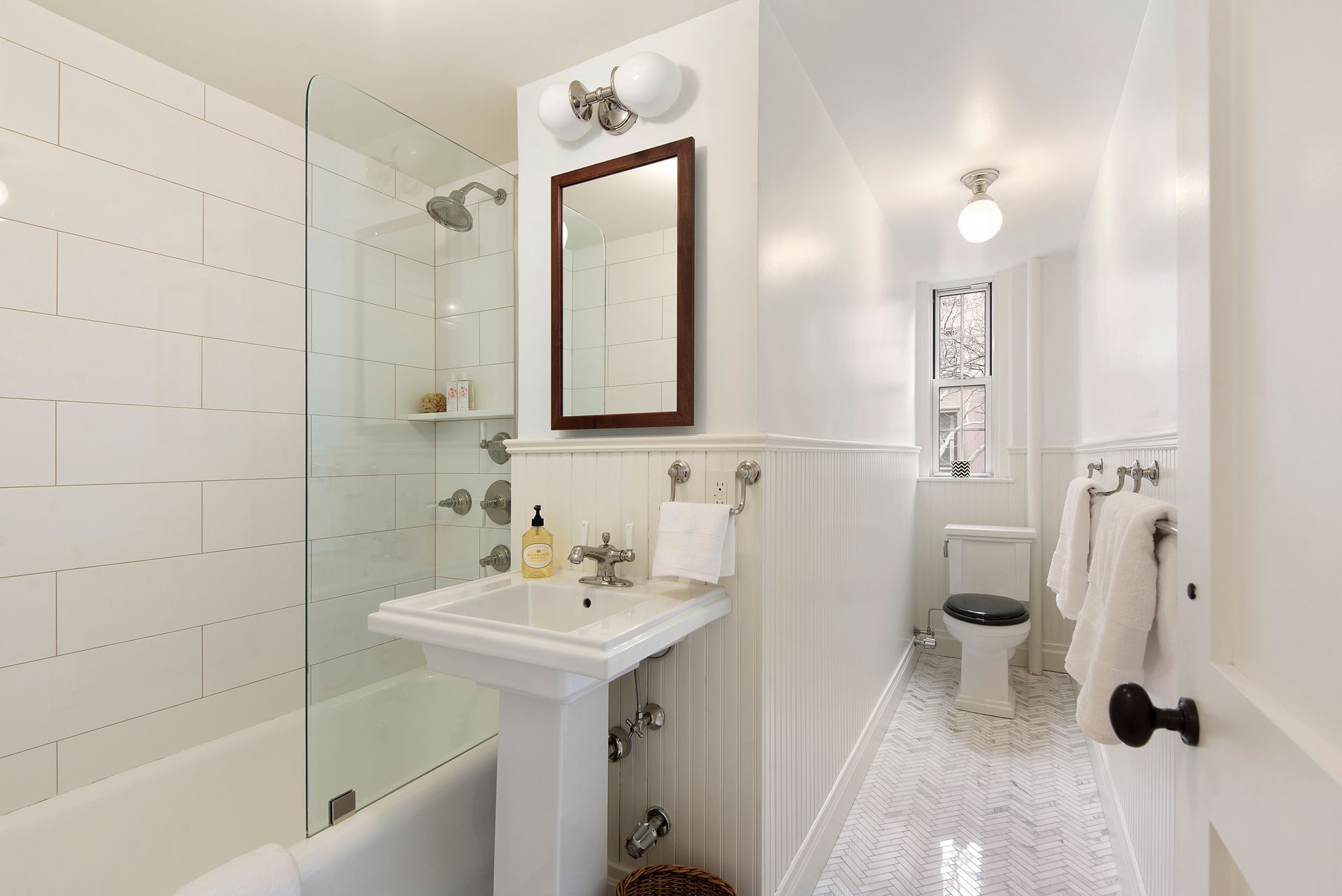 A bright, narrow white bathroom with a pedestal sink, glass-enclosed bathtub, toilet, and marble-patterned tile floor.
