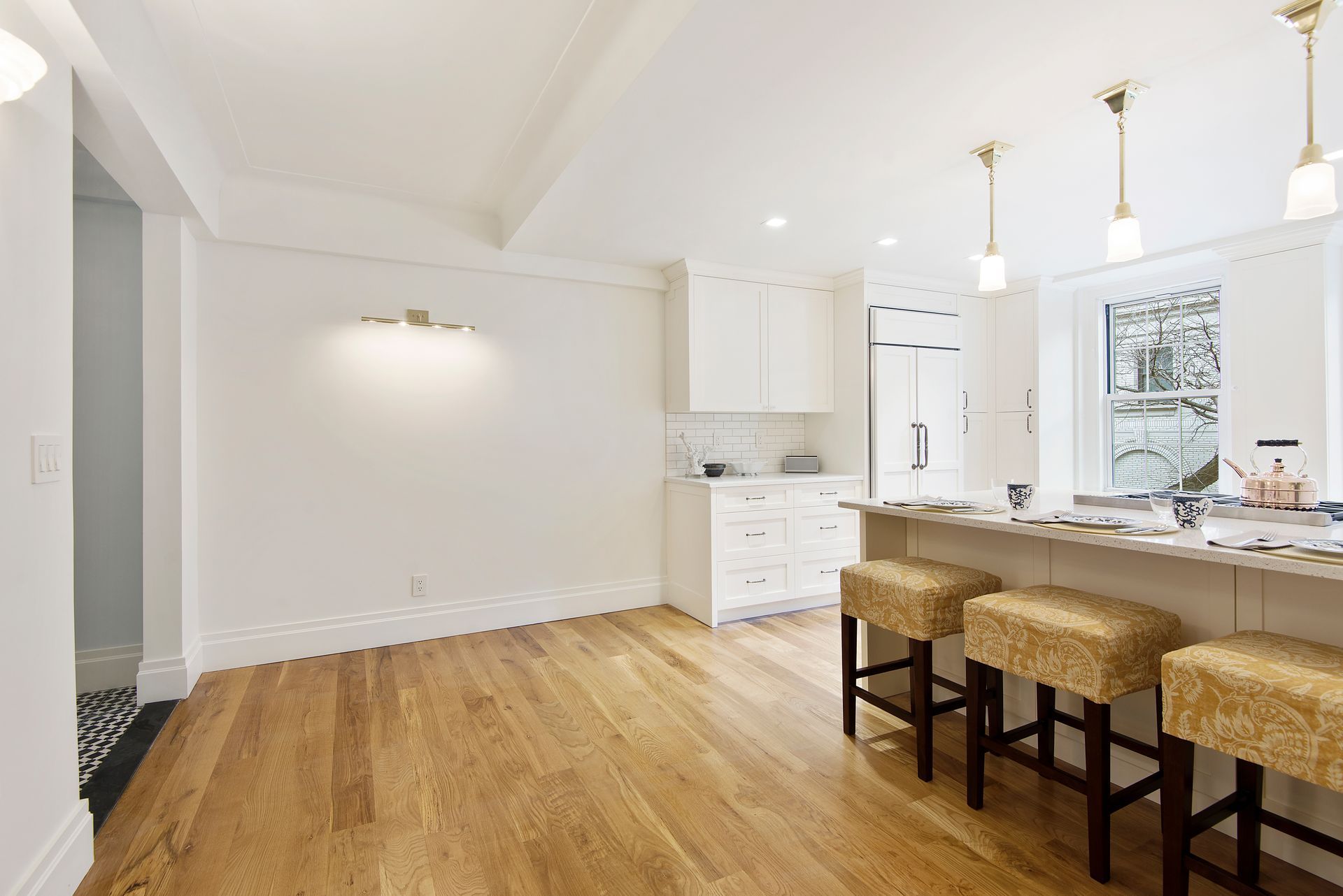 A bright kitchen with white cabinetry, light wood floors, and a breakfast bar with three textured stools.