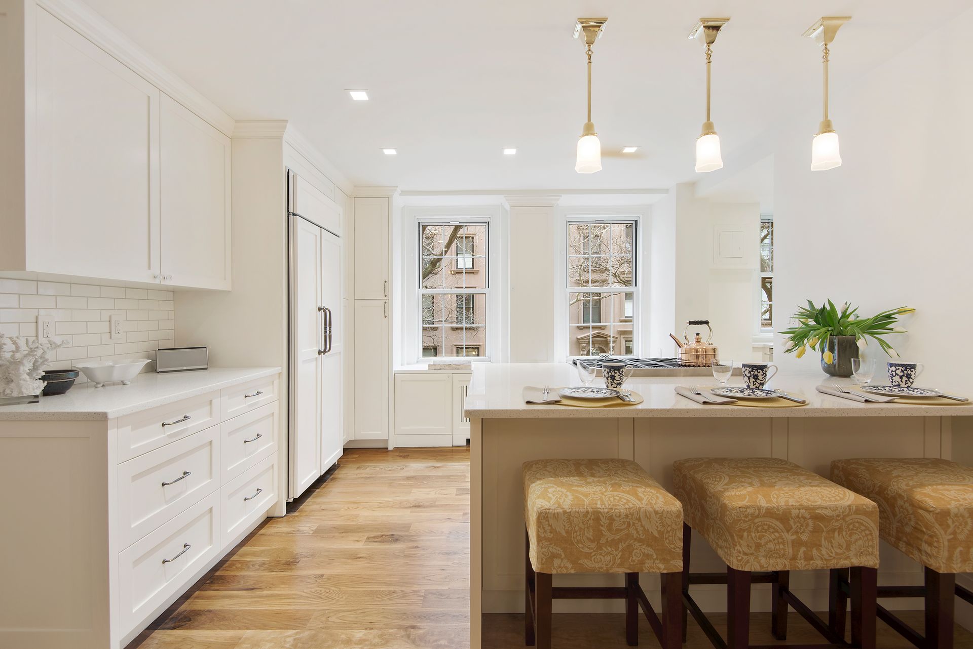 A bright, modern kitchen featuring white cabinets, a long island with three beige stools, and light wood flooring.