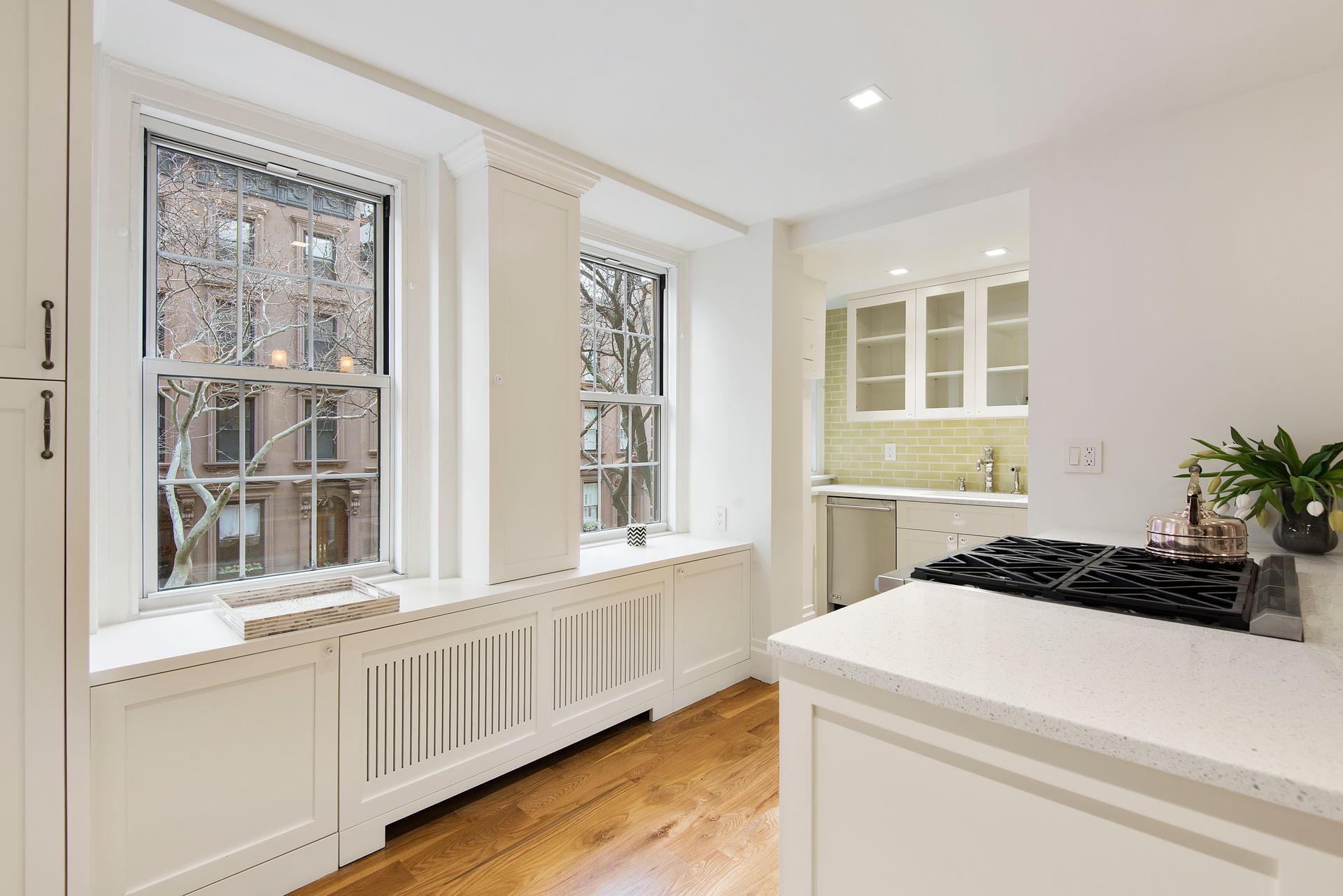 A bright kitchen with white cabinets, a light countertop, wood flooring, and two windows with white radiator covers.