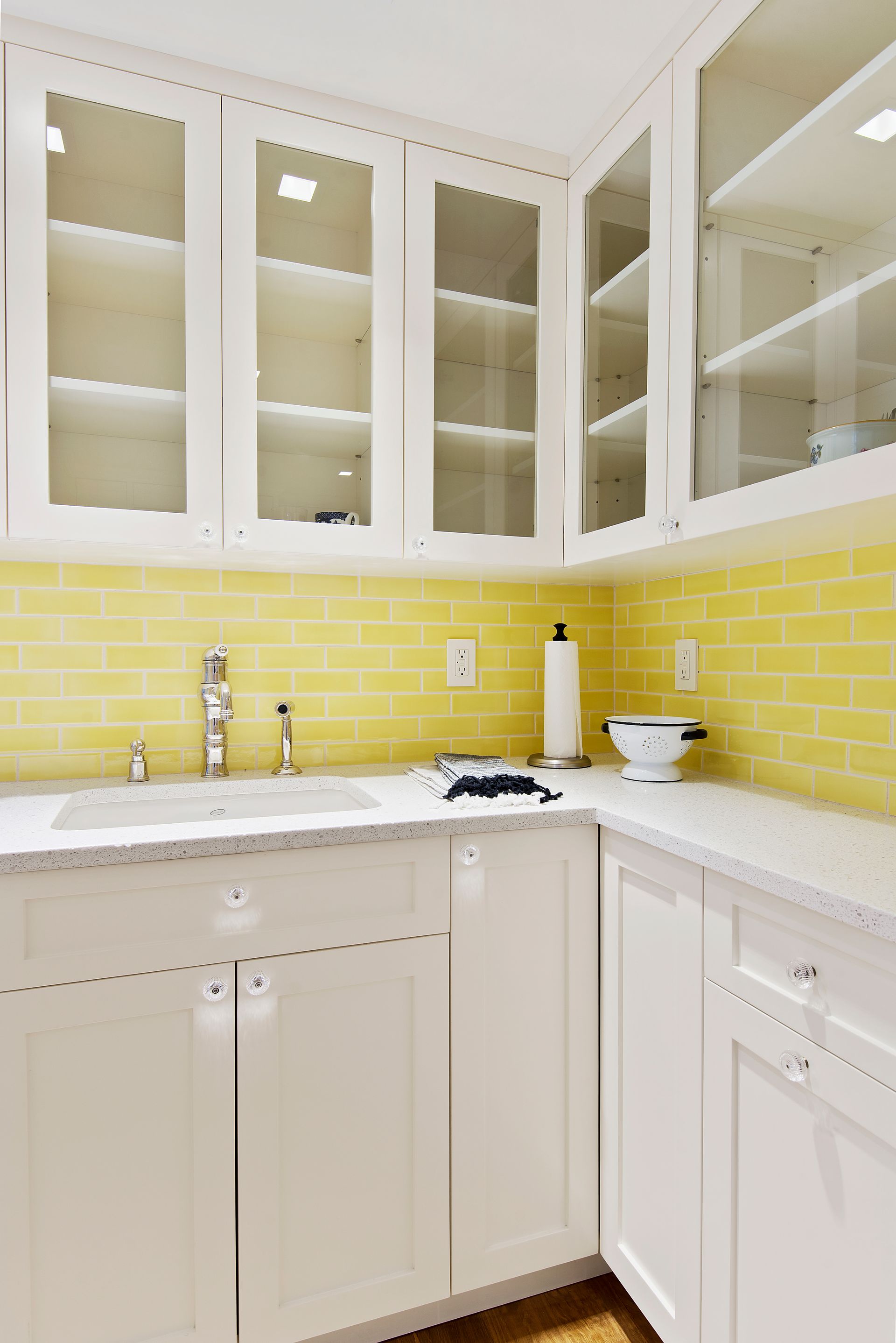 Kitchen sink area with white cabinets, glass-front upper cupboards, and a bright yellow subway tile backsplash.