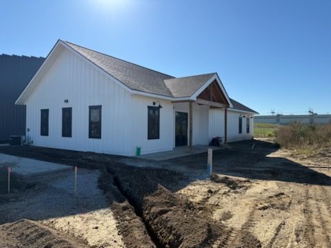 White farmhouse under construction, dark roof and windows, dirt trenches in front, bright sunlight.