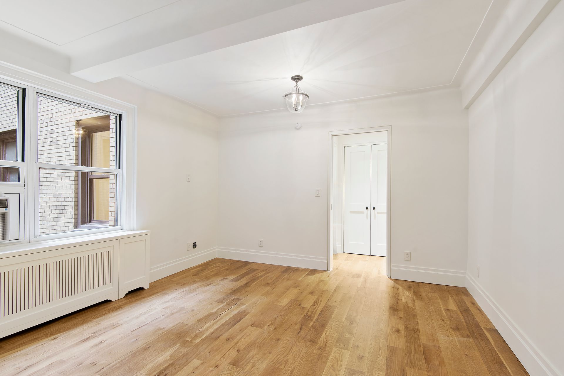 An empty room with white walls, light wood floors, a radiator under a window, and a doorway leading to a hallway.