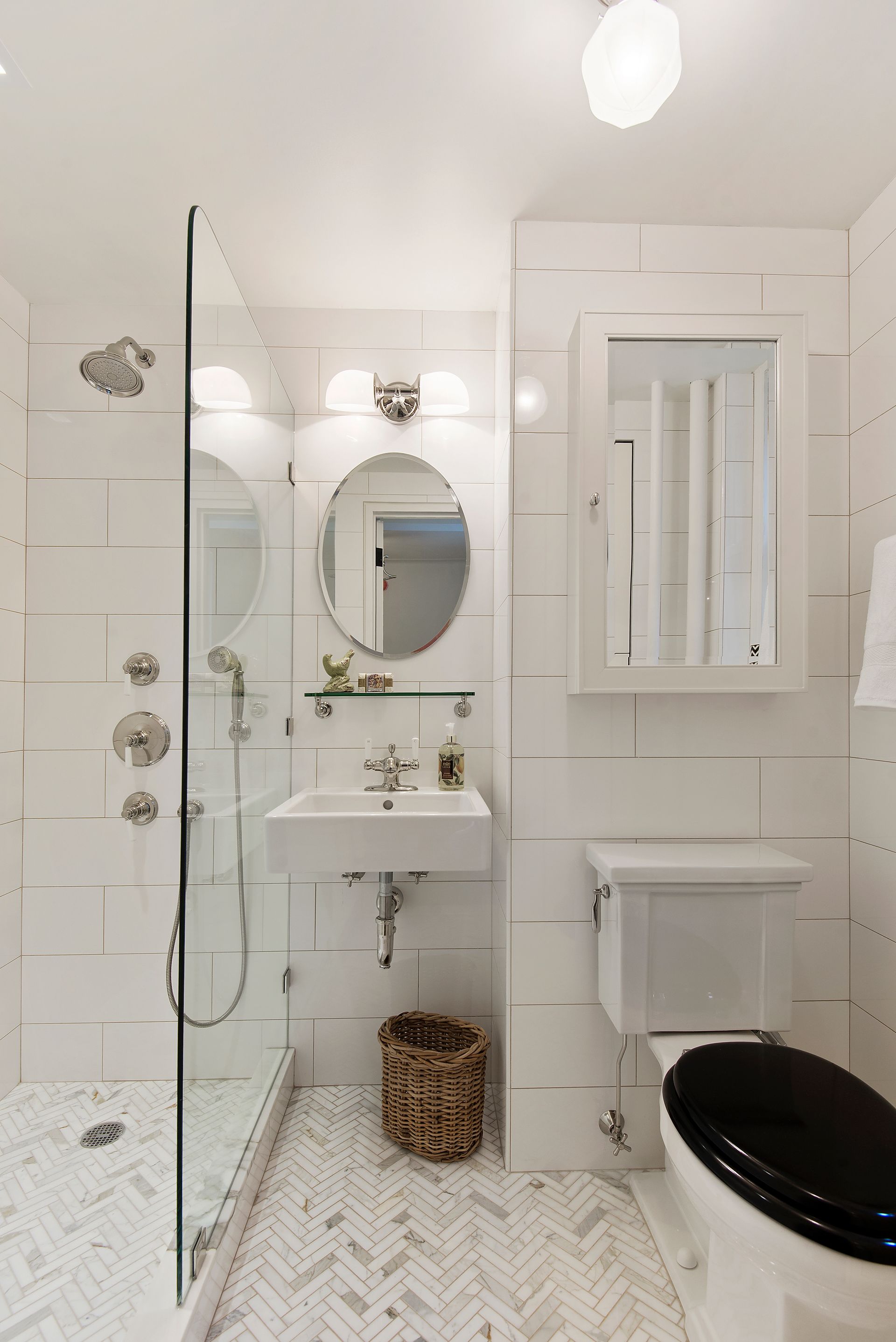 Modern bathroom with a glass-enclosed shower, wall-mounted sink, oval mirror, white tiled walls, and a black toilet seat.