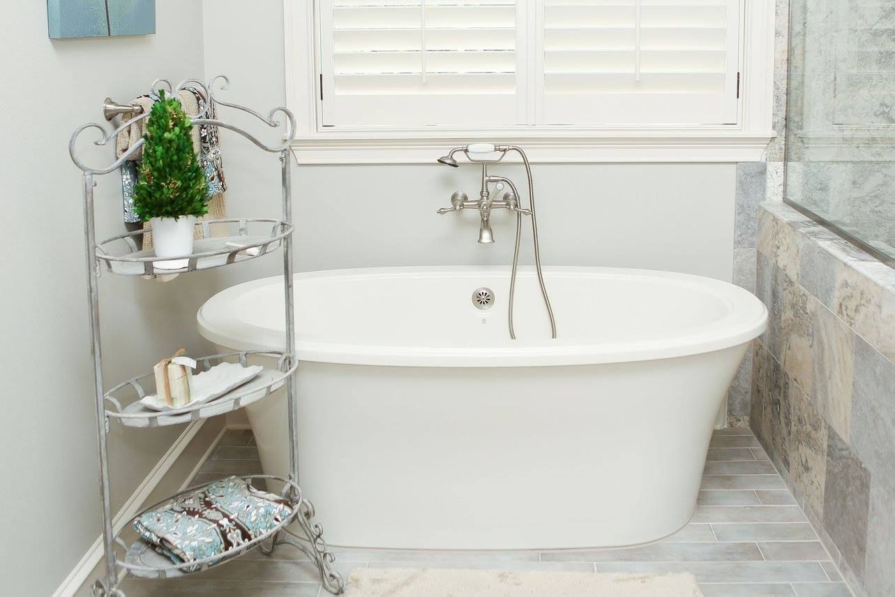 White oval bathtub in a light blue bathroom, with a metal shelf, window with shutters, and gray stone flooring.