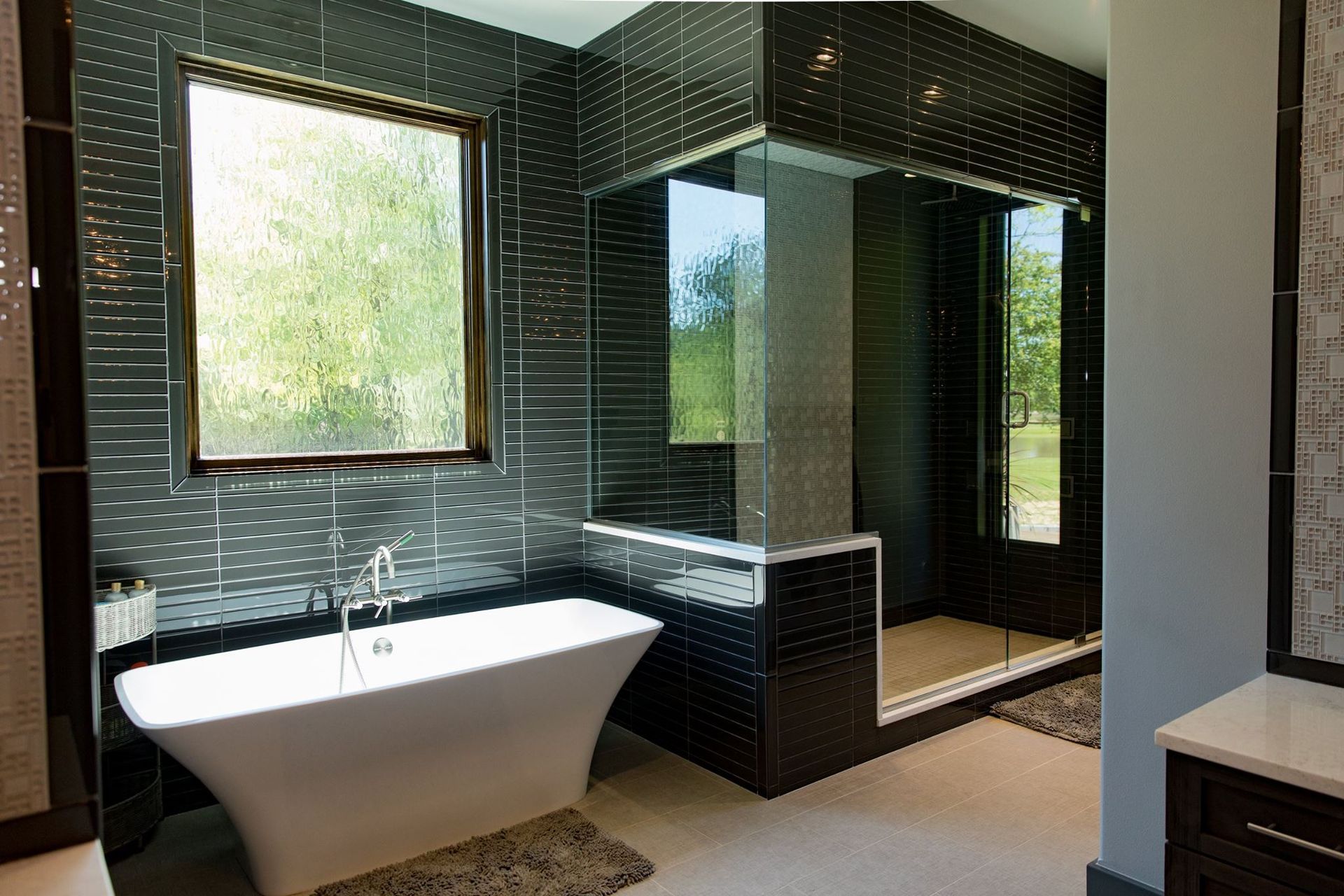 Modern bathroom with black tiled walls, a white tub, glass shower, and large window.