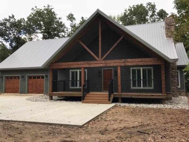 Gray cabin-style house with metal roof, wooden porch, and attached garage. Brown doors and trim.