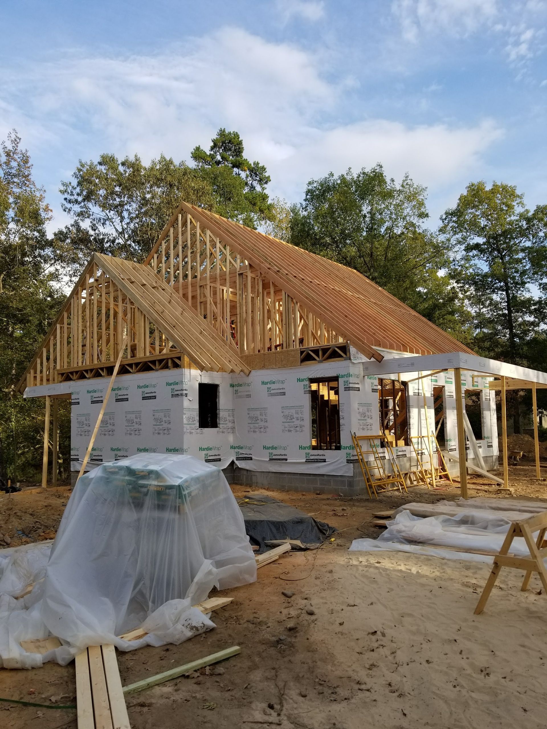 House under construction, wooden frame with roof trusses. Wrapped in white sheathing with trees in the background.